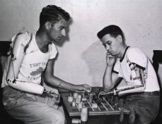 Two men, each with a prosthetic arm, sit at a table and play checkers.