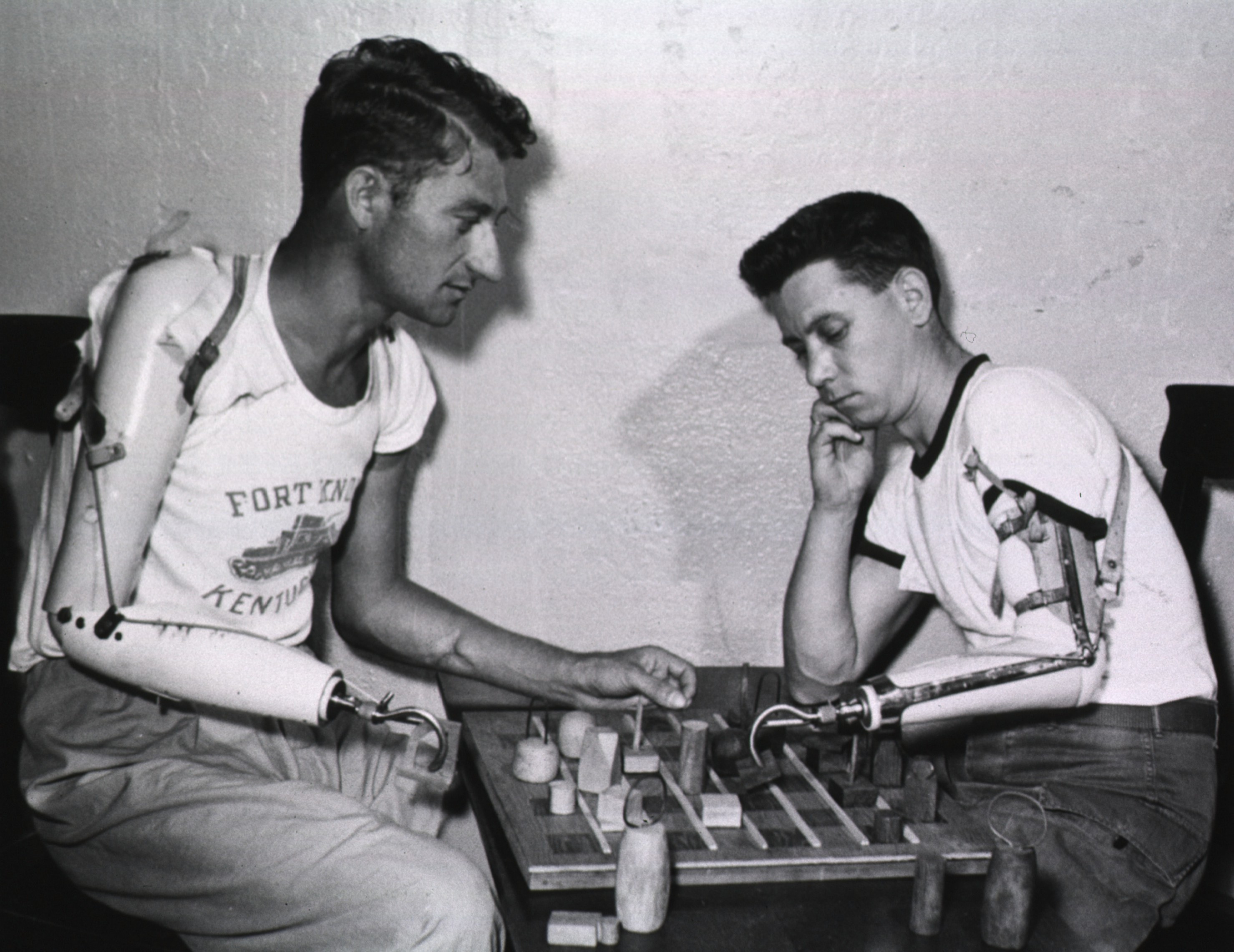 Two men, each with a prosthetic arm, sit at a table and play checkers. 